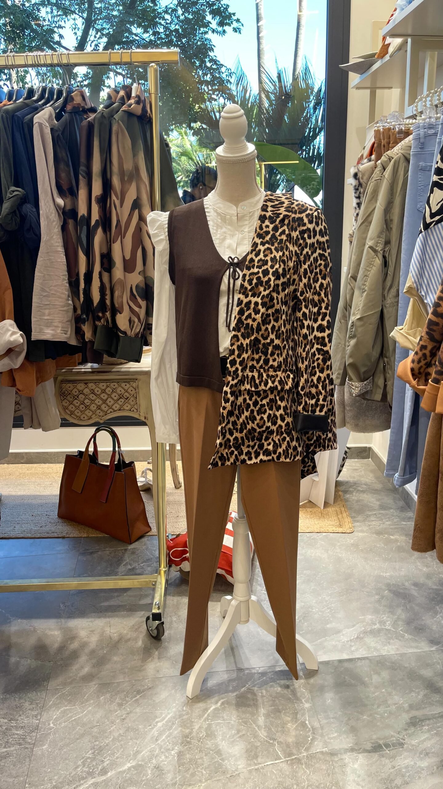 Maniquí con un conjunto de pantalones marrones, camisa blanca y chaleco marrón, con una chaqueta de estampado de leopardo colgada sobre un hombro. Ropa en perchas detrás.