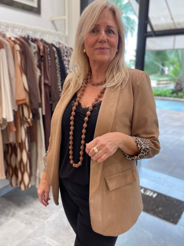 Mujer con blazer beige y puños de estampado animal, una blusa negra y un collar largo de cuentas marrones, en el interior de una tienda de ropa con estantes llenos de prendas en tonos neutros de fondo.