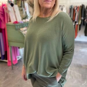 Mujer rubia con un conjunto de jersey y pantalones verde oliva. La escena de fondo muestra ropa y decoraciones en tonos coloridos.