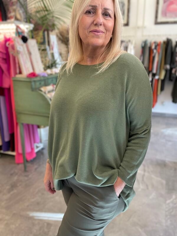 Mujer rubia con un conjunto de jersey y pantalones verde oliva. La escena de fondo muestra ropa y decoraciones en tonos coloridos.