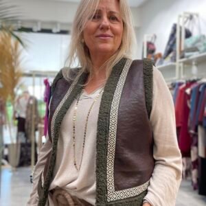 Mujer con chaleco marrón oscuro con ribetes de lana verde y detalles en patrones geométricos blancos. Lleva un cinturón ancho marrón y un collar largo de cuentas doradas.