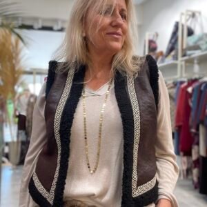Mujer con el mismo chaleco marrón oscuro, esta vez con ribetes de lana negra y detalles geométricos blancos. Lleva un collar dorado y un cinturón ancho.