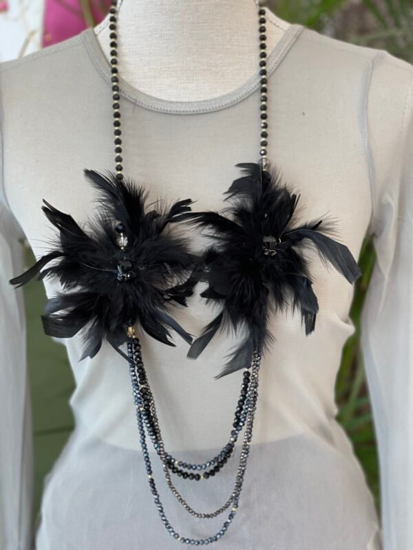 Collar decorativo con cuentas y dos grandes adornos de plumas negras, exhibido en un busto de tela gris claro. El collar tiene varias cadenas colgantes de cuentas negras y plateadas.