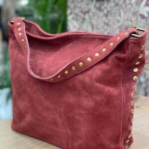 Bolso grande de gamuza color rojizo, con detalles de tachuelas doradas en el asa y los laterales, sobre una superficie de madera clara.