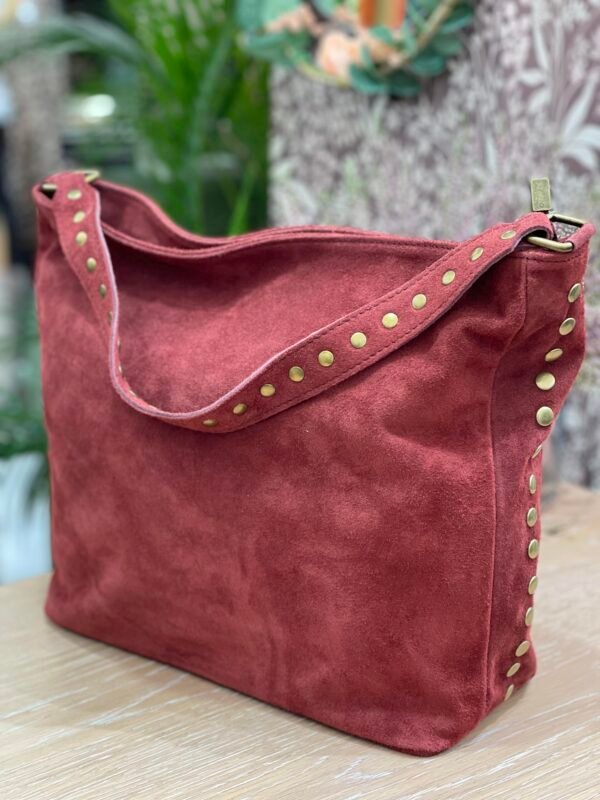 Bolso grande de gamuza color rojizo, con detalles de tachuelas doradas en el asa y los laterales, sobre una superficie de madera clara.