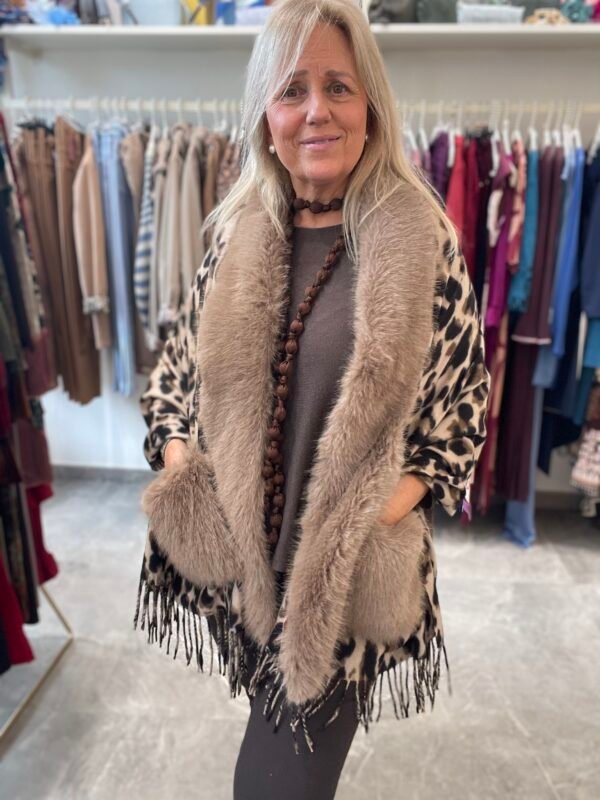 Mujer con un abrigo de estampado de leopardo y cuello de peluche beige. Lleva un collar de cuentas marrones y ropa oscura, con un fondo de ropa en exhibición.