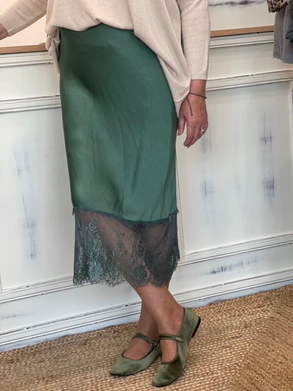 Mujer con falda verde satinada con encaje en el dobladillo y zapatos tipo Mary Jane en terciopelo verde, sobre una alfombra de yute.