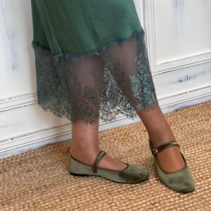 Detalle de falda verde satinada con encaje en el borde inferior y zapatos Mary Jane de terciopelo verde, sobre una alfombra de yute.