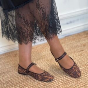 Detalle de una falda negra satinada con encaje floral en el borde inferior. La modelo lleva zapatos planos tipo Mary Jane con estampado de leopardo, sobre una alfombra de yute.