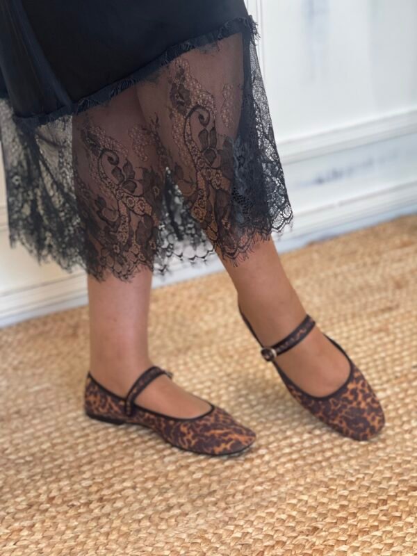 Detalle de una falda negra satinada con encaje floral en el borde inferior. La modelo lleva zapatos planos tipo Mary Jane con estampado de leopardo, sobre una alfombra de yute.