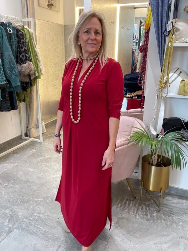 Mujer con vestido largo rojo de manga tres cuartos, acompañado de un collar largo de cuentas beige, en el interior de una tienda de ropa.