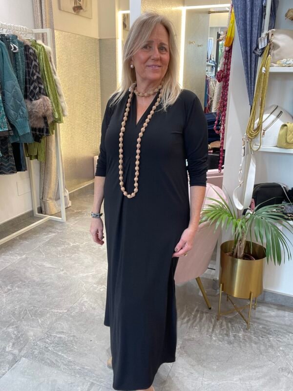 Mujer con vestido largo negro de manga tres cuartos, usando un collar largo de cuentas beige, en el interior de una tienda de ropa.